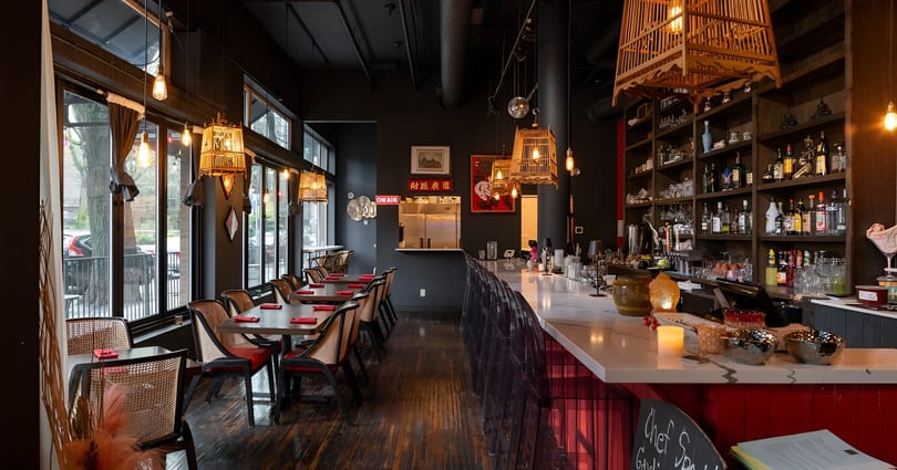 Restaurant interior with dark walls, wooden tables, wicker chairs, a long bar, and decorative lighting.