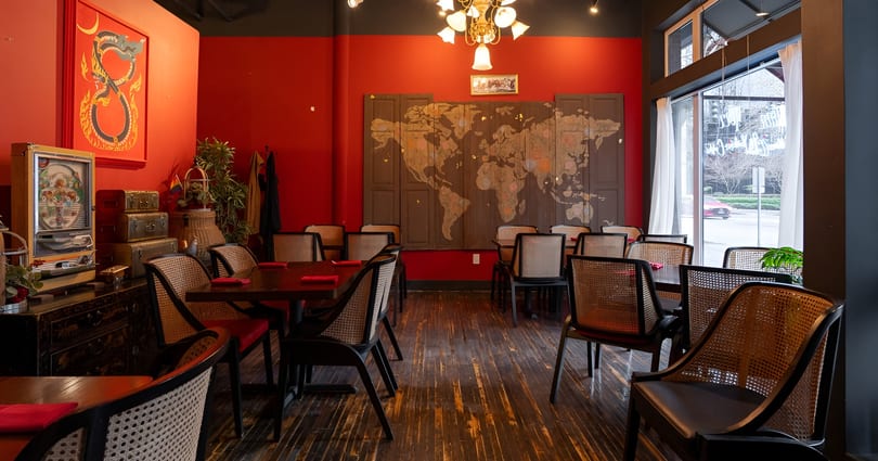 Restaurant dining room with red walls, dark wood floors, cane-back chairs, and a world map mural.