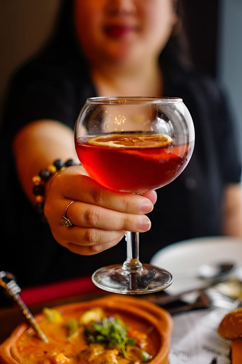 A person holding a glass of red cocktail with an orange slice, with food in the foreground.