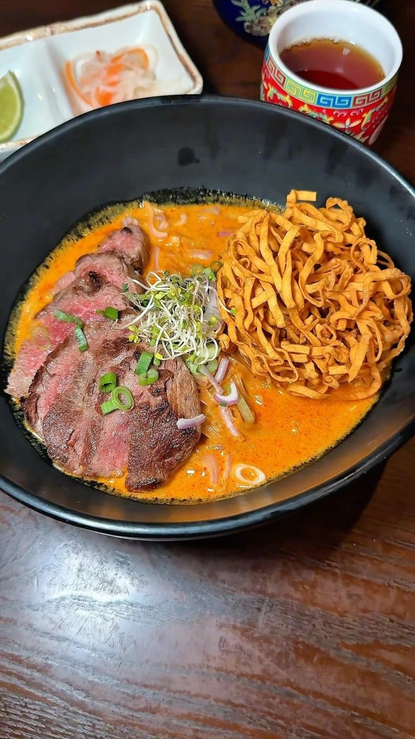 Khao Soi with sliced beef, crispy noodles, sprouts, and green onions in a black bowl.
