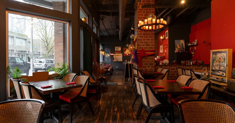 Restaurant interior with dark wood tables, wicker chairs, red walls, and a large window overlooking a street.