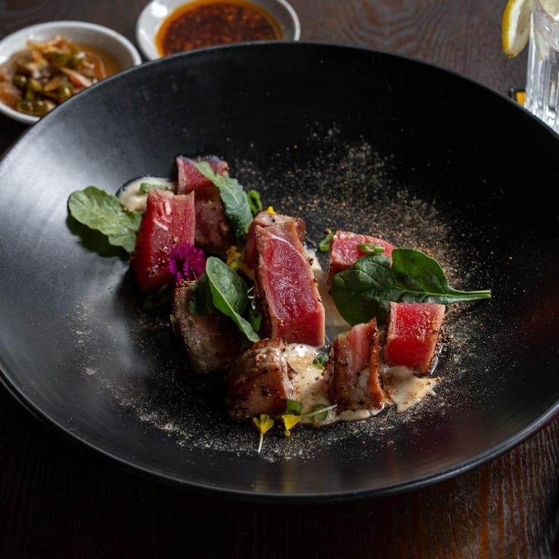 Seared tuna slices with greens, sauce, and spices on a black plate, with side dishes.