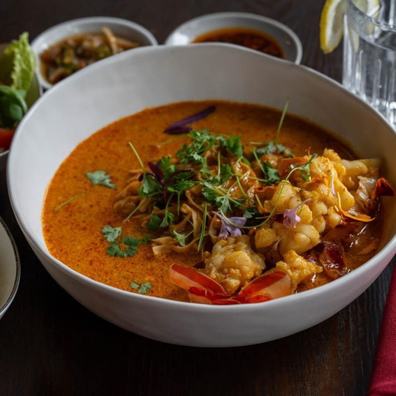 A white bowl filled with a vibrant orange curry, shrimp, noodles, and fresh herbs.
