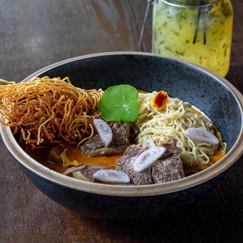 Khao Soi with beef, crispy noodles, fresh noodles, shallots, and a green leaf in a dark bowl.