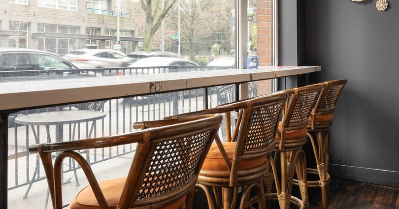 Rattan bar stools with orange cushions line a window counter overlooking a city street.