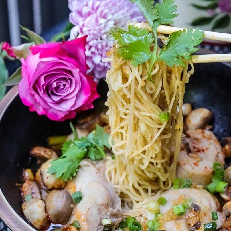 Chopsticks lifting noodles from a bowl of chicken, mushrooms, and green onions, with flowers.