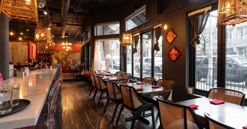 Interior of a restaurant with a long bar, dining tables, and large windows looking out onto a street.