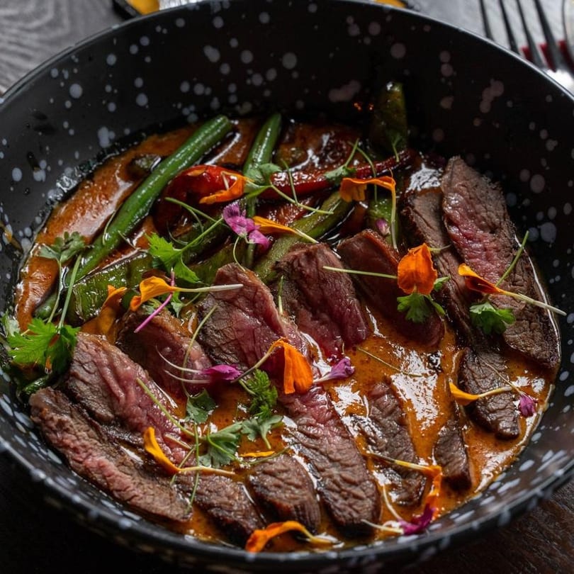 Sliced steak in a rich orange sauce with green beans, red peppers, and edible flowers.