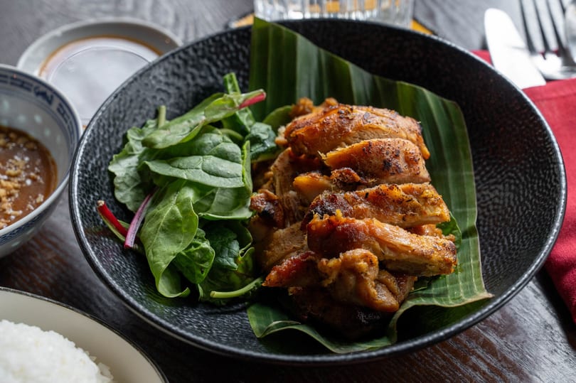 Grilled chicken slices on a banana leaf with a side of fresh spinach in a black bowl.